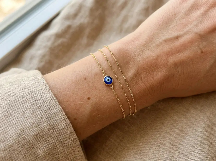 Wrist wearing a slim bracelet stack with one tiny blue evil eye charm between plain fine chains.