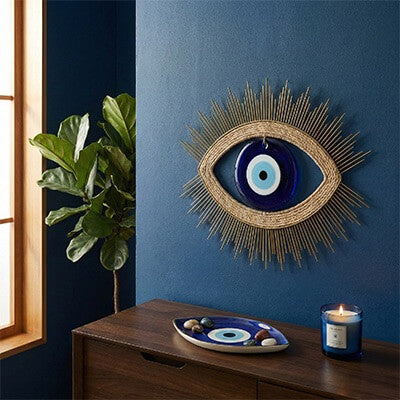 Stylish interior scene showcasing curated Evil Eye home decor, including a large woven and blue glass wall hanging above a wooden console with a ceramic eye tray and lit candle.