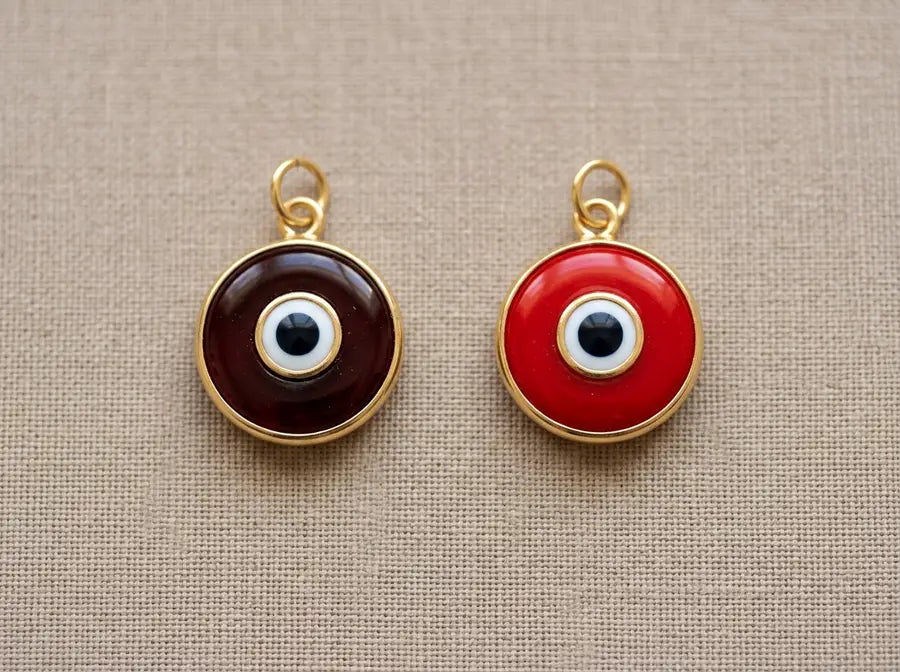 A deep oxblood-red evil eye pendant beside a bright scarlet version of the same pendant on a warm neutral surface