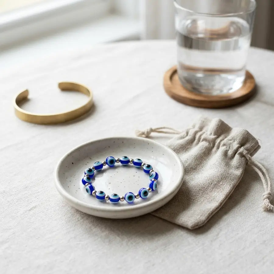 Finished blue evil eye bracelet rests in a ceramic dish beside a fabric pouch, water glass, and metal bangle.