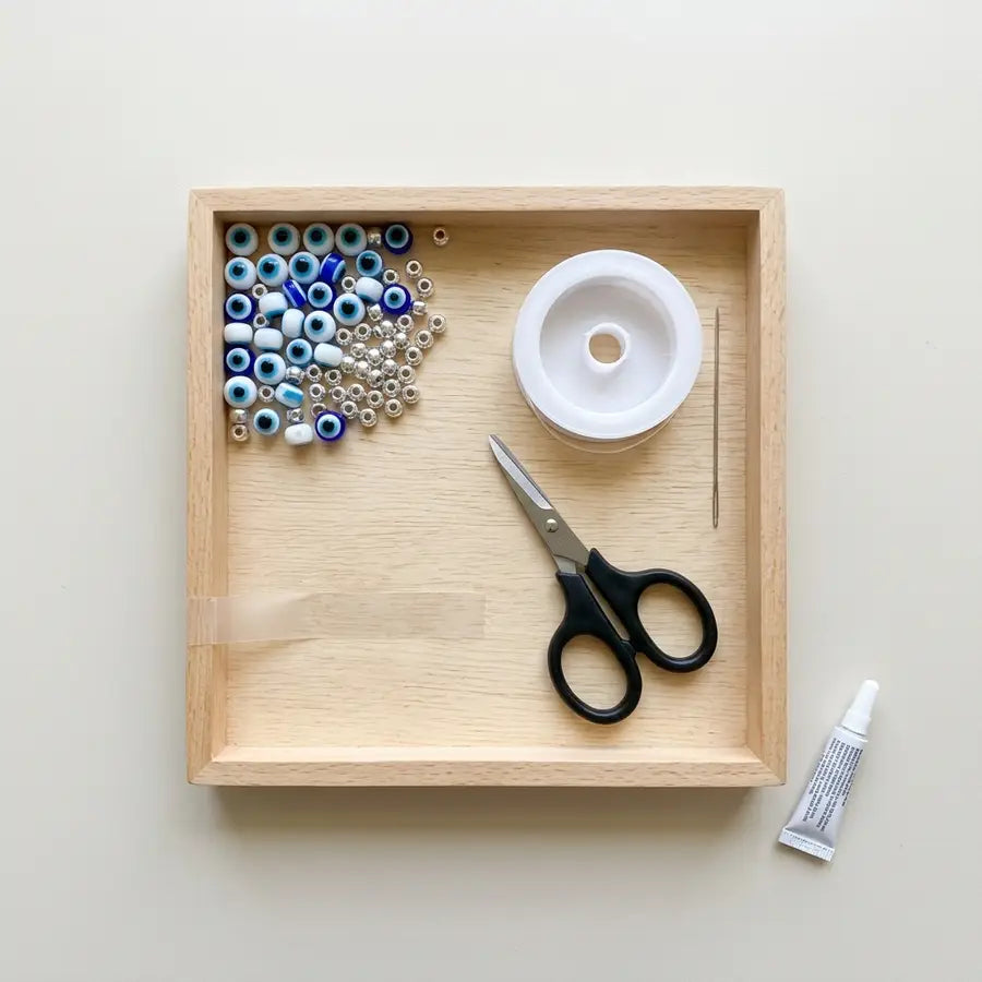 Shallow tray with evil eye beads, spacers, stretch cord spool, scissors, tape, needle, and small glue tube spaced clearly.