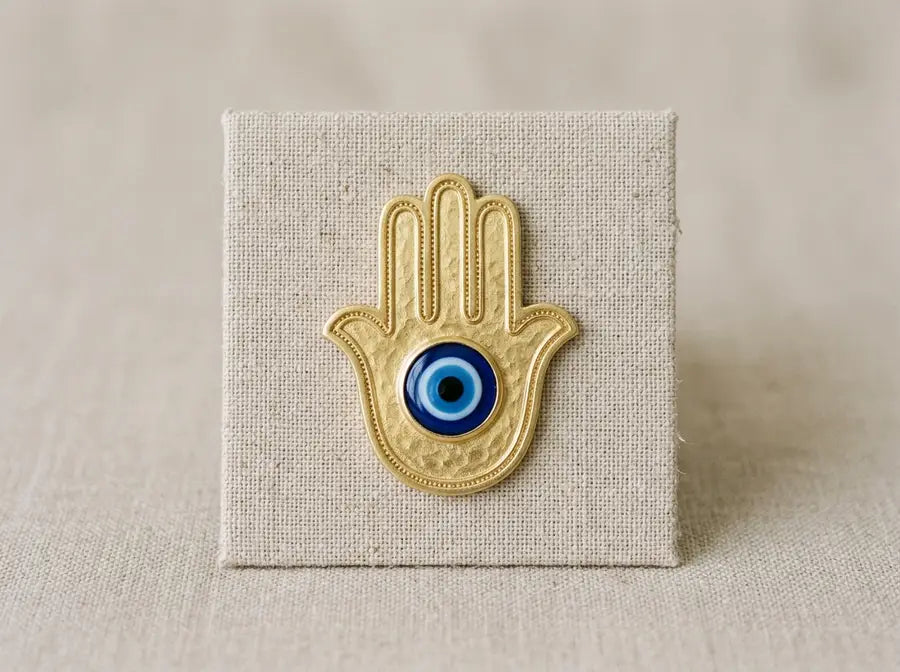 Flat gold Hamsa palm with five spread fingers and a blue central eye laid on a plain linen card