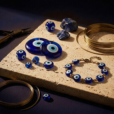 A detailed still life showing the craftsmanship behind Evil Eye jewelry, featuring loose blue glass beads, lapis lazuli stones, brass wire, and tools arranged on a textured stone slab under focused warm light.