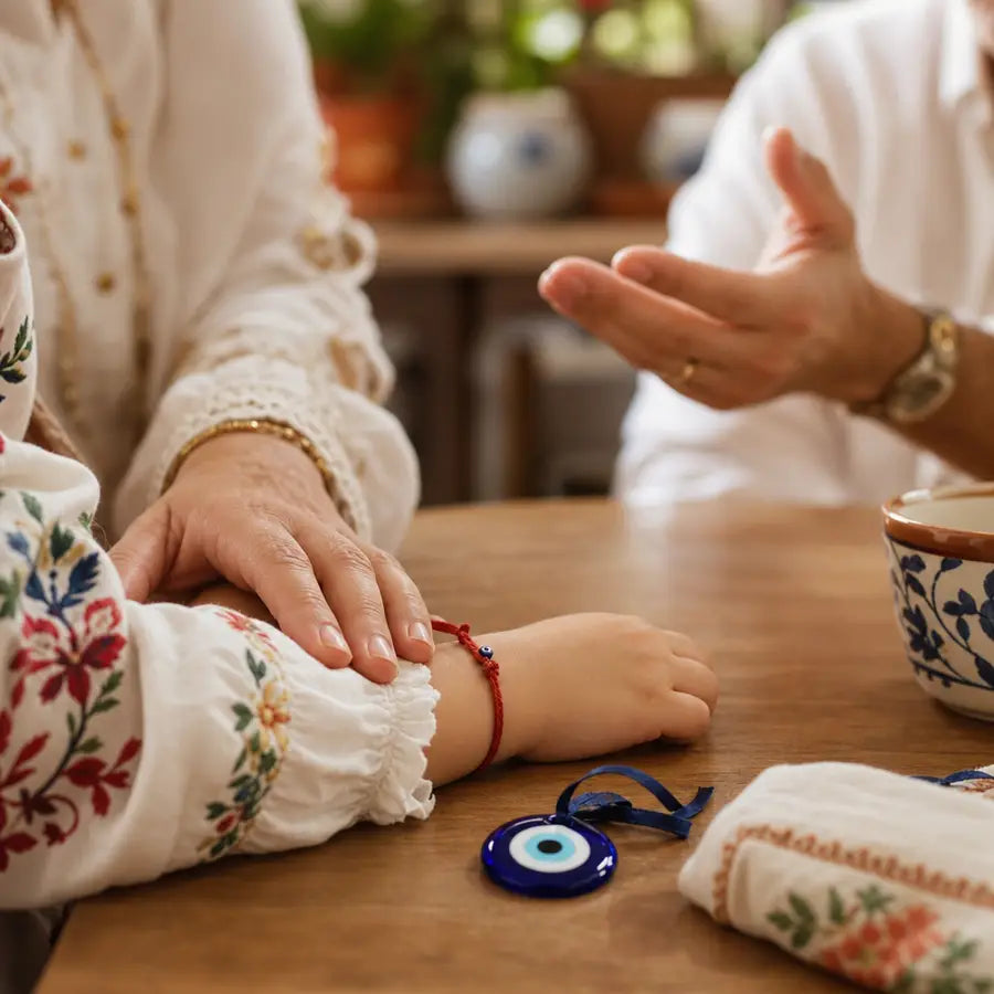 Adult hand gently touching a child’s embroidered sleeve and red bracelet, with a blue evil eye charm between the child and a