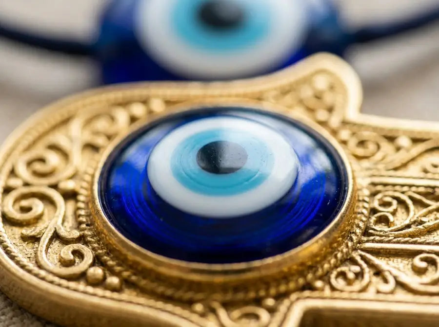Macro close-up of a blue concentric eye in a gold Hamsa palm with a blurred blue Evil Eye bead behind it