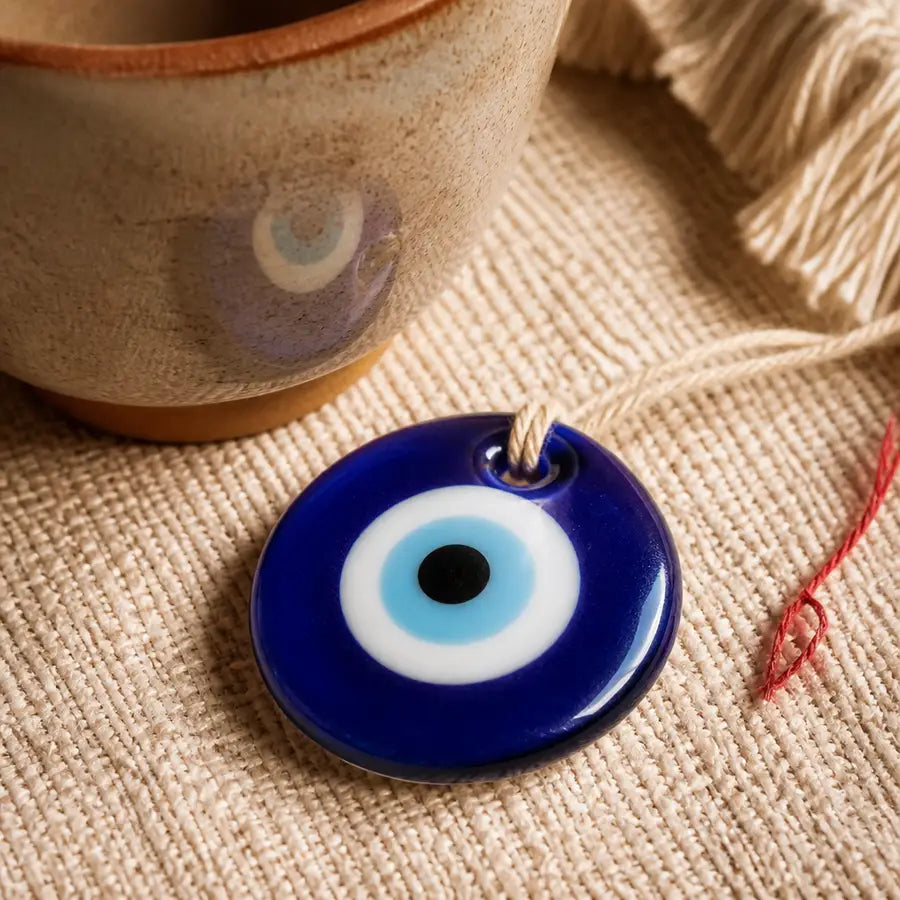 Close-up blue evil eye pendant on woven cloth with a faint gaze-like reflection on a ceramic cup nearby