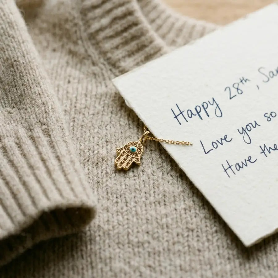 Small gold Hamsa pendant beside a fine chain, handwritten birthday note, and oatmeal knit sleeve suggesting everyday wear.