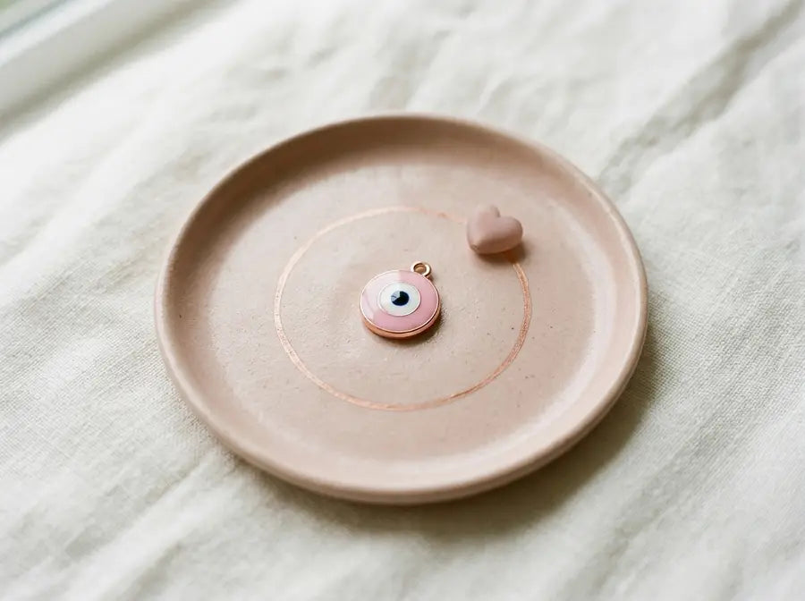 Single pink evil eye charm inside a rose-gold circle with a small heart bead on a blush ceramic tray