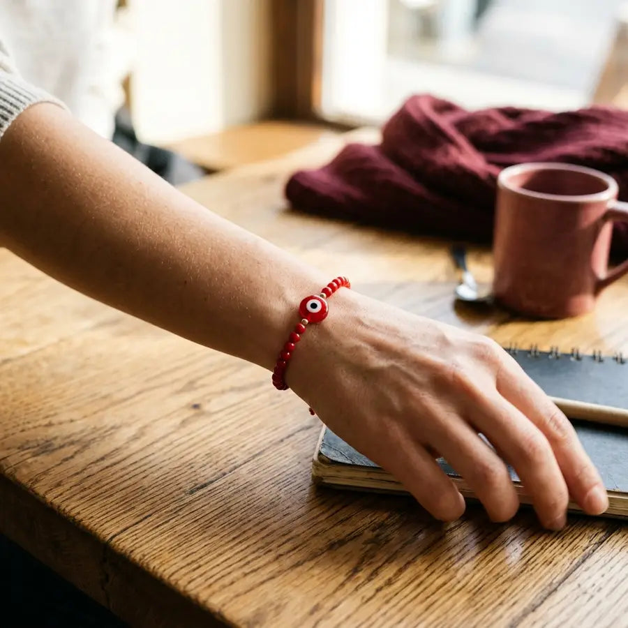 Red evil eye bracelet on a moving wrist above a wooden table with a blurred maroon scarf behind it.