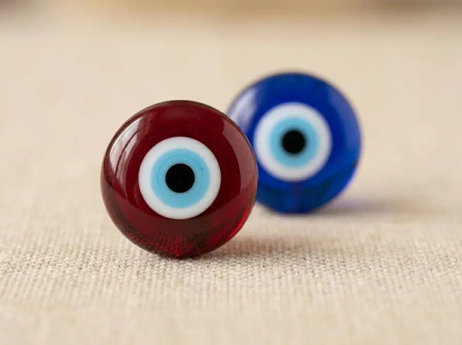A red evil eye bead and a blue evil eye bead of identical shape placed side by side on a warm neutral surface