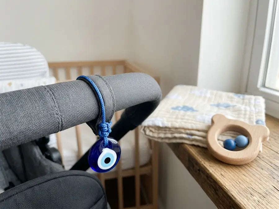 Small blue evil eye charm fastened to the outside loop of a stroller handle, away from the baby area, with a folded blanket n