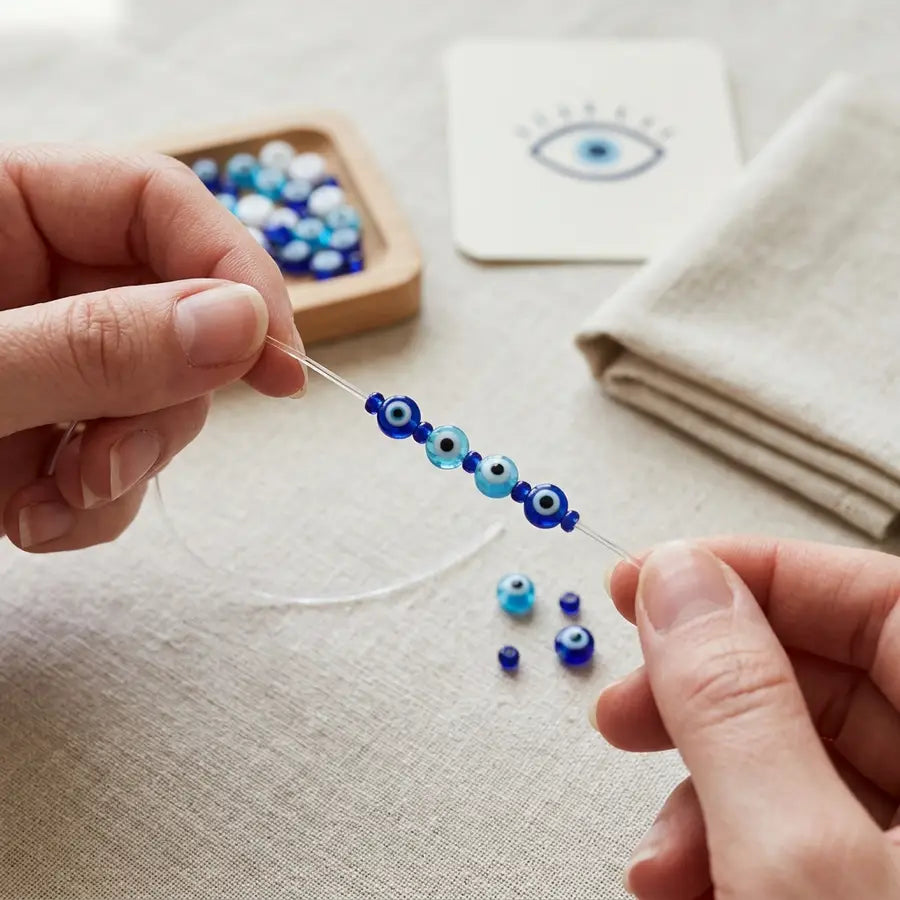 Sharp foreground stretch cord and partially strung evil eye bracelet with a blurred blue eye motif card behind it.
