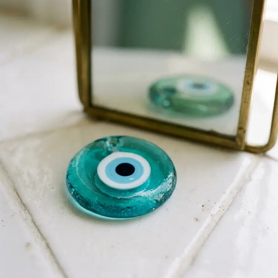 Sharp turquoise evil eye charm in front with a softer greener reflection behind it on white ceramic tile.