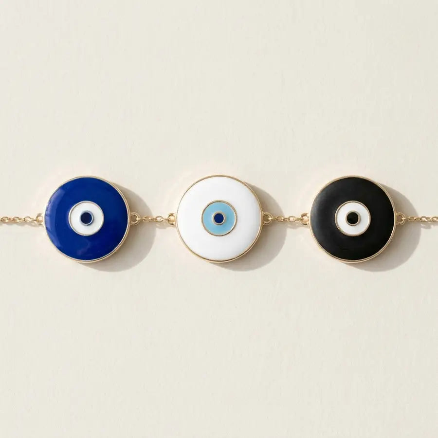 Three same-size evil eye charms in blue, white, and black aligned on ivory, with the white charm brightest in the center