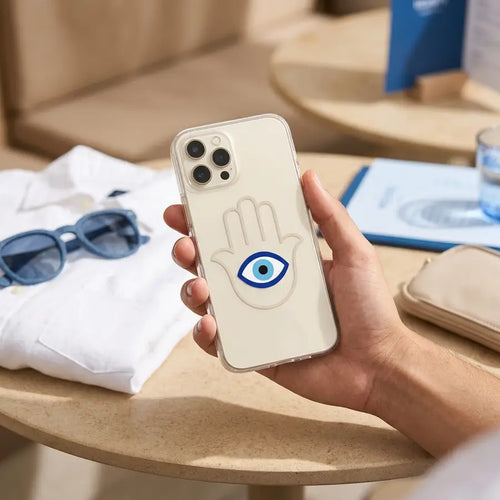 Clear phone case with a blue eye inside a Hamsa outline, held above sunglasses and travel items.