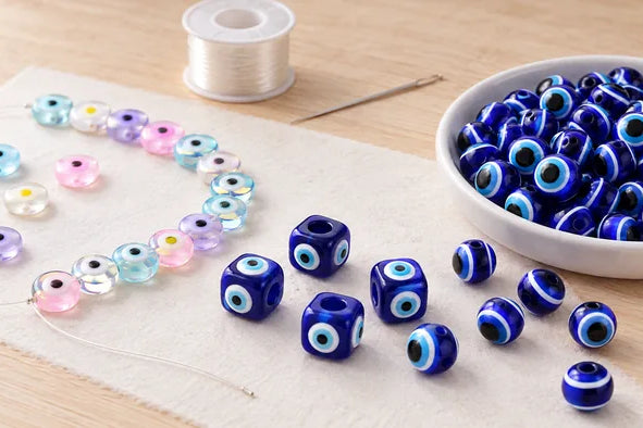 Blue and pastel evil eye beads arranged on a craft mat beside a bowl of beads and thread
