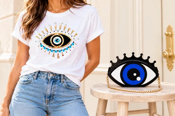 Woman wearing a white evil eye T-shirt beside an eye-shaped bag on a pale wooden stool