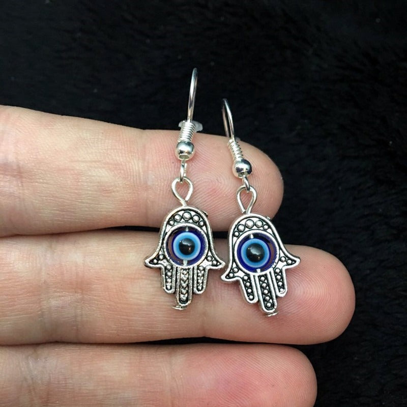 Silver Hamsa hand earrings with blue eyes held between fingers against a dark background