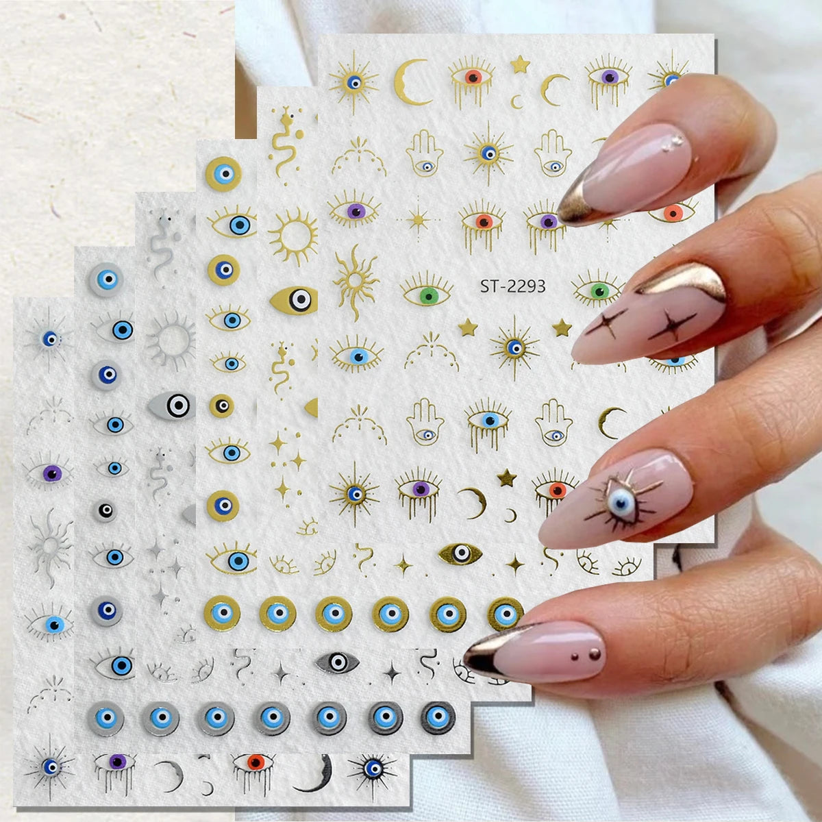Nail art sticker sheets with various eye, crescent moon, and hamsa designs, held by a hand with manicured nails against a light fabric background.