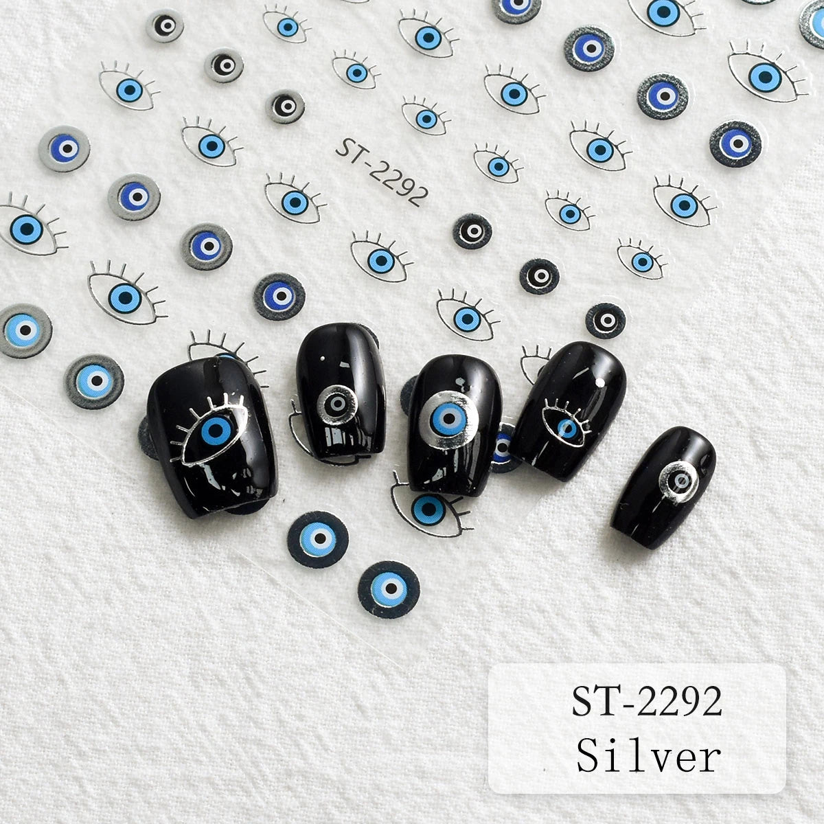 Five black nail wraps decorated with evil eye designs are arranged on a textured white background, alongside loose matching evil eye stickers and printed sample illustrations. The text 'ST-2292 Silver' appears in the bottom right corner.