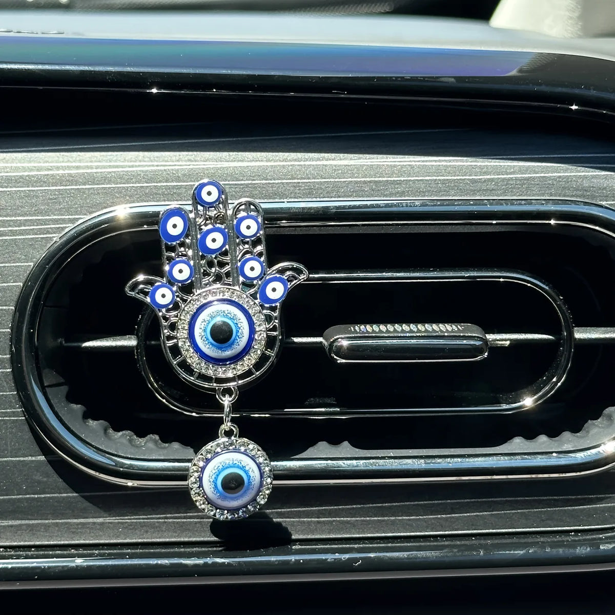 One silver tone Hamsa car vent clip with blue eye motifs, clear accents and a round hanging eye pendant.