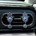 Front view of two silver tone Hamsa clips on a dark car vent, showing open hand outlines and clear accents around the blue eyes.