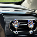 Two gold tone Hamsa clips displayed across a car vent, each with blue eye motifs and a round eye pendant below.
