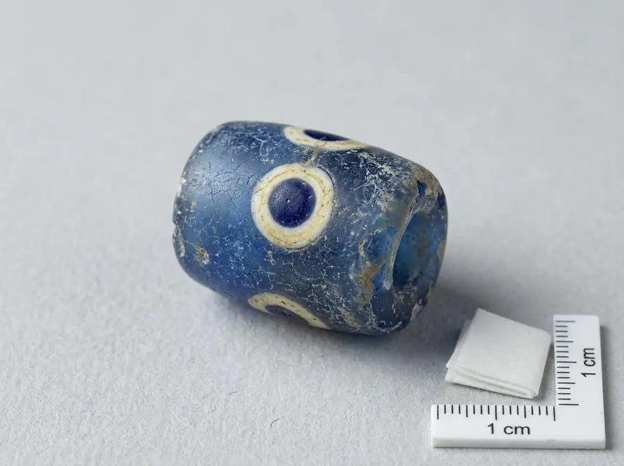 A weathered cylindrical blue glass bead with applied white-and-blue eye spots rests on archival board beside a small scale ma
