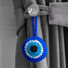 Blue round evil eye charm suspended by its matching loop from a clear button on a grey curtain tieback.