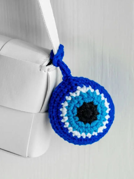 Blue round evil eye charm looped around a white bag strap, with a black center and turquoise blue and white rings.