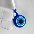 Blue round evil eye charm looped around a white bag strap, with a black center and turquoise blue and white rings.