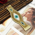 Gold tone beaded cuff bracelet with three eye motifs, laid diagonally beside bamboo on a magazine.