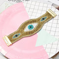 Gold tone beaded cuff bracelet laid open on pink and grid paper, showing three eye motifs and both end fittings.