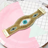 Gold tone beaded cuff bracelet laid open on pink and grid paper, showing three eye motifs and both end fittings.