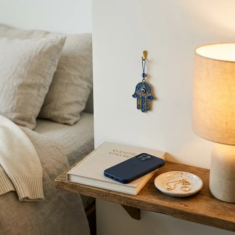 Small Hamsa charm on a wall hook above a bedside shelf with face-down phone, closed book, jewelry dish, lamp, and folded card