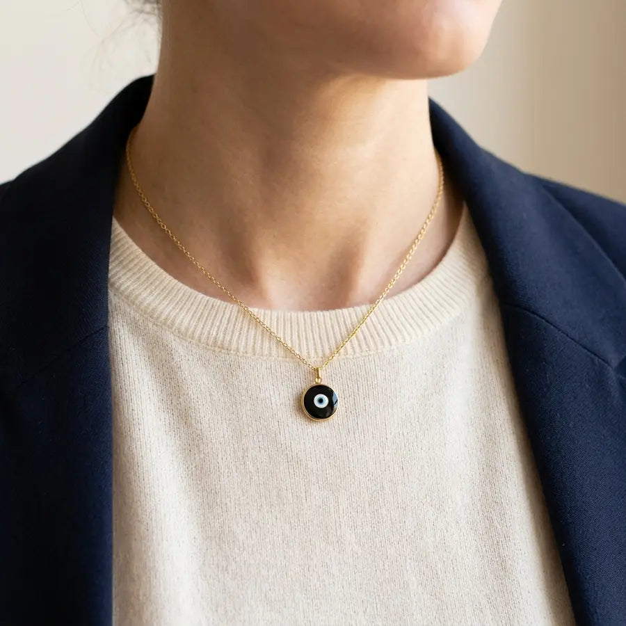 Black evil eye pendant on a fine gold chain resting below the collarbone against an ivory neckline and navy blazer.