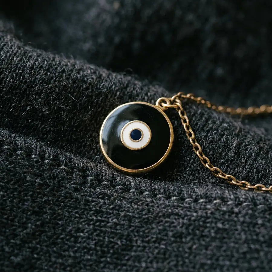 Close-up black enamel evil eye charm on charcoal knit fabric with a gold chain and crisp textured shadows.