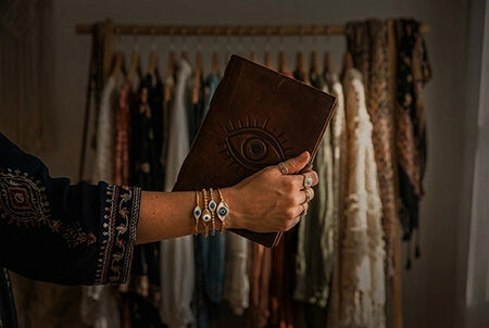 A fashion shot showing an arm with a dark embroidered sleeve and layered evil eye bracelets, holding a mystical leather journal against a dimly lit bohemian clothing rack.