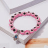 Pink eye bead bracelet with white rings, black centers and a dangling silver tone Hamsa hand with a blue eye, resting on an open book.