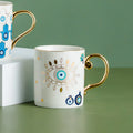 White coffee cup with a turquoise eye, gold tone leaf border and curved gold tone handle.