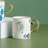 White coffee cup with a turquoise eye, gold tone leaf border and curved gold tone handle.
