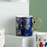 Navy blue coffee cup with gold tone lotus flowers, Namaste lettering, Hamsa motifs and a curved gold tone handle.