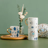Three white coffee cups with blue Hamsa and eye patterns and gold tone handles, arranged beside flowers and an eye-shaped plate.