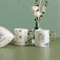 Two white coffee cups with scattered eye and leaf border eye designs, each with a gold tone handle.