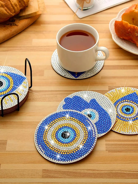 Blue and gold tone eye and Hamsa coasters arranged on a wooden table with a black holder and a white cup on one coaster.