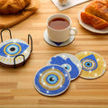 Blue and gold tone eye and Hamsa coasters arranged on a wooden table with a black holder and a white cup on one coaster.