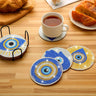 Blue and gold tone eye and Hamsa coasters arranged on a wooden table with a black holder and a white cup on one coaster.