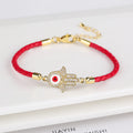 Red braided bracelet with a gold tone Hamsa centerpiece, clear accents, and a red eye on white