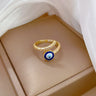 Gold tone blue evil eye bypass ring resting on a taupe display cushion