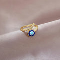 Gold tone open bypass ring with a round blue evil eye and clear accents, resting on taupe fabric